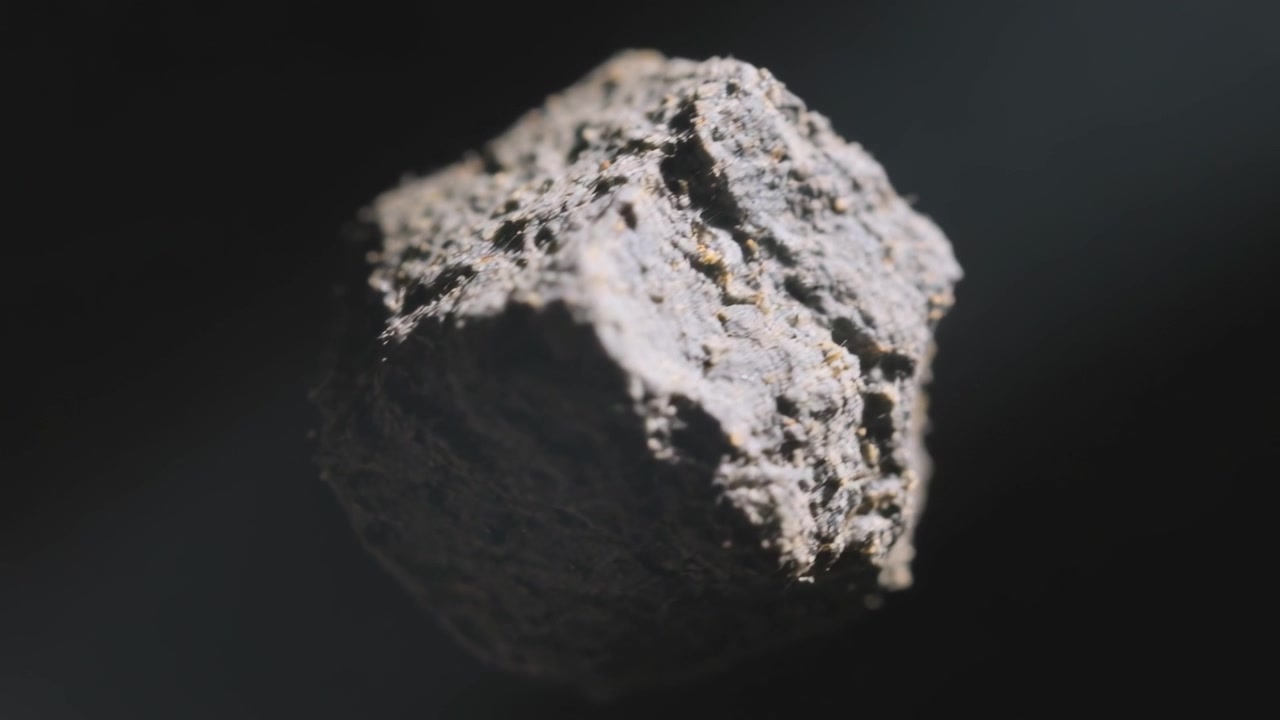 Rotating after camera start, dark-gray pellet revealing textured faces in studio, with gold flecks