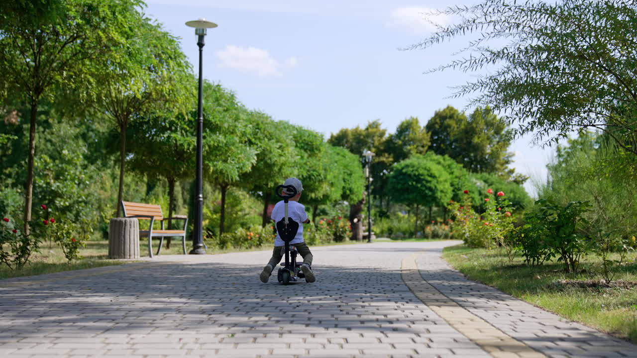 Rear view of a toddler distancing from a camera on his bike. Kid pushes from the ground to move by the paved road in the park.