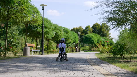 rear view of a toddler distancing from a camera on his bike. kid pushes from the ground to move by the paved road in the park.
