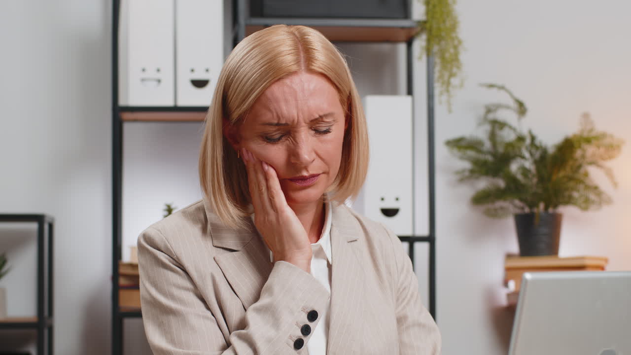 Upset mature businesswoman in suit suffering from toothache while using laptop at home office desk