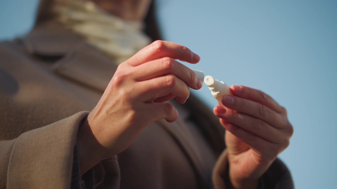 Primo piano di donna che tiene e apre una piccola bottiglia bianca all'aperto sotto un cielo azzurro e limpido. Mani con unghie smaltate manipolano l'oggetto, enfatizzando l'interazione tra mani e prodotto.