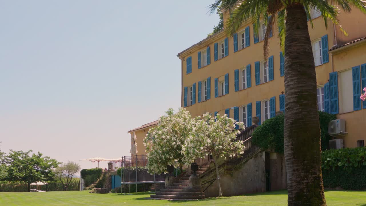 A wide shot captures a charming yellow building with blue shutters in Italy, surrounded by a lush lawn, palm trees, and blooming white flowers under a clear sky.