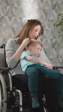 little girl reads book while mom holding toddler in arms sitting in wheelchair. young woman with disability spends evening with children at home