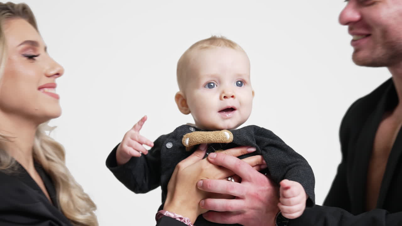 Parents hold their beloved tiny baby boy from two sides. Loving mom and dad kiss their child on the head. White backdrop.