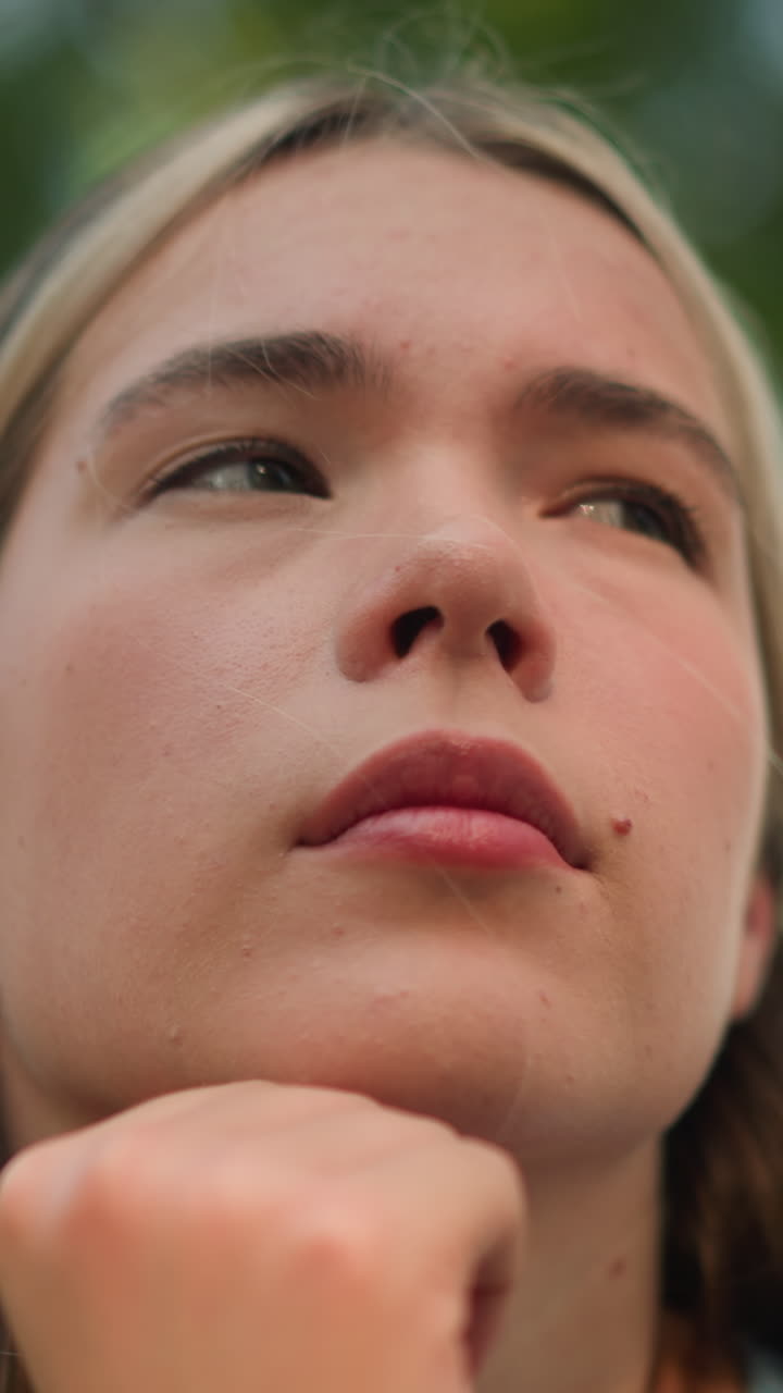 Close-up of young woman with hand under chin, looking pensive while glancing into distance, expressing deep thought blurred greenery in the background, sunlight softly illuminates her face