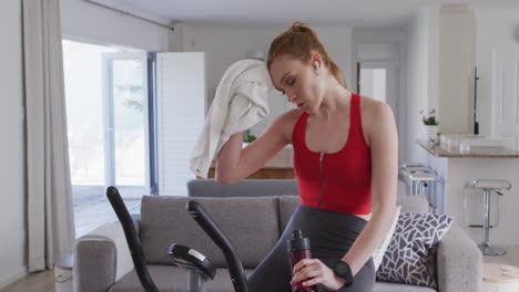 Woman-drinking-water-and-wiping-her-face-with-a-towel-while-sitting-on-stationary-bike-at-home