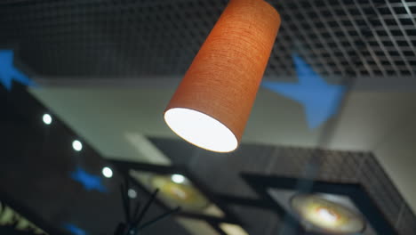 vista de cerca de una lámpara colgante iluminada con una sombra naranja, colgando del techo con un fondo de estrellas azules y un techo con patrón de cuadrícula