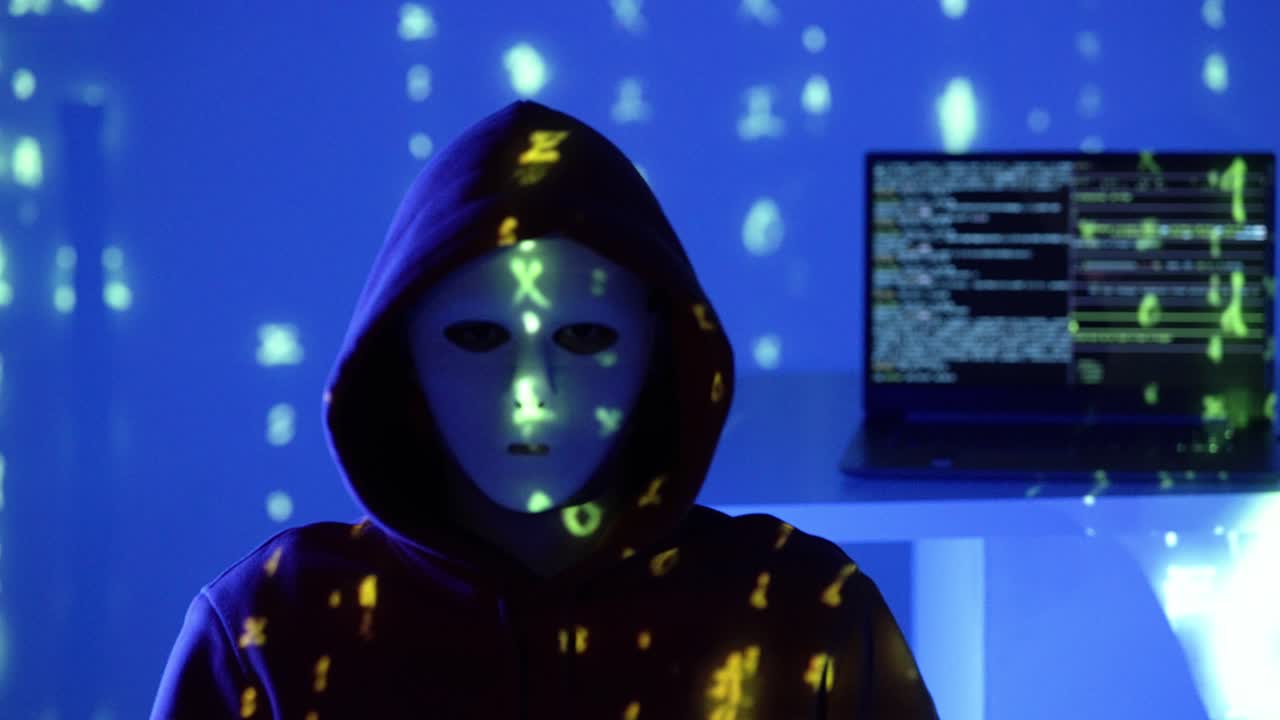 Masked individual in a dark hoodie sits in front of a glowing laptop, surrounded by cryptic blue code projections, evoking cyber infiltration, stealth hacking, and digital espionage under secrecy
