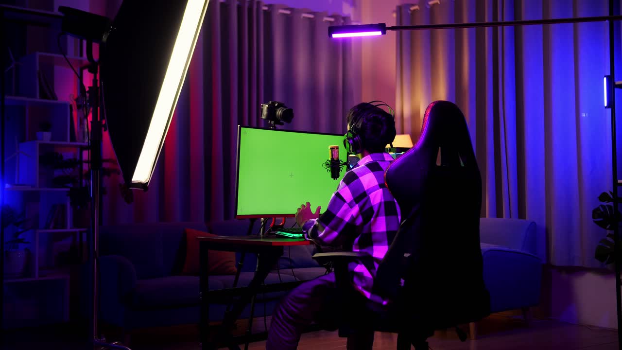 Back View Of Asian Boy Streamer Using Green Screen Personal Computer, Live Stream Waving Hand And Talking On The Microphone.  Desk Illuminated By Rgb Led Strip Light