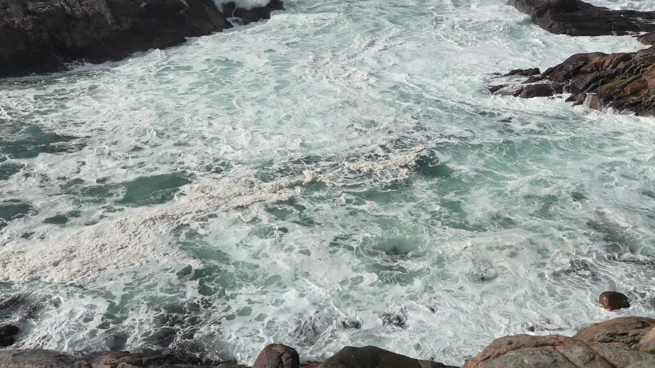 Foamy waves tumble over rugged rocks along the coastline during a bright sunny day. The clear blue water contrasts with dark stones