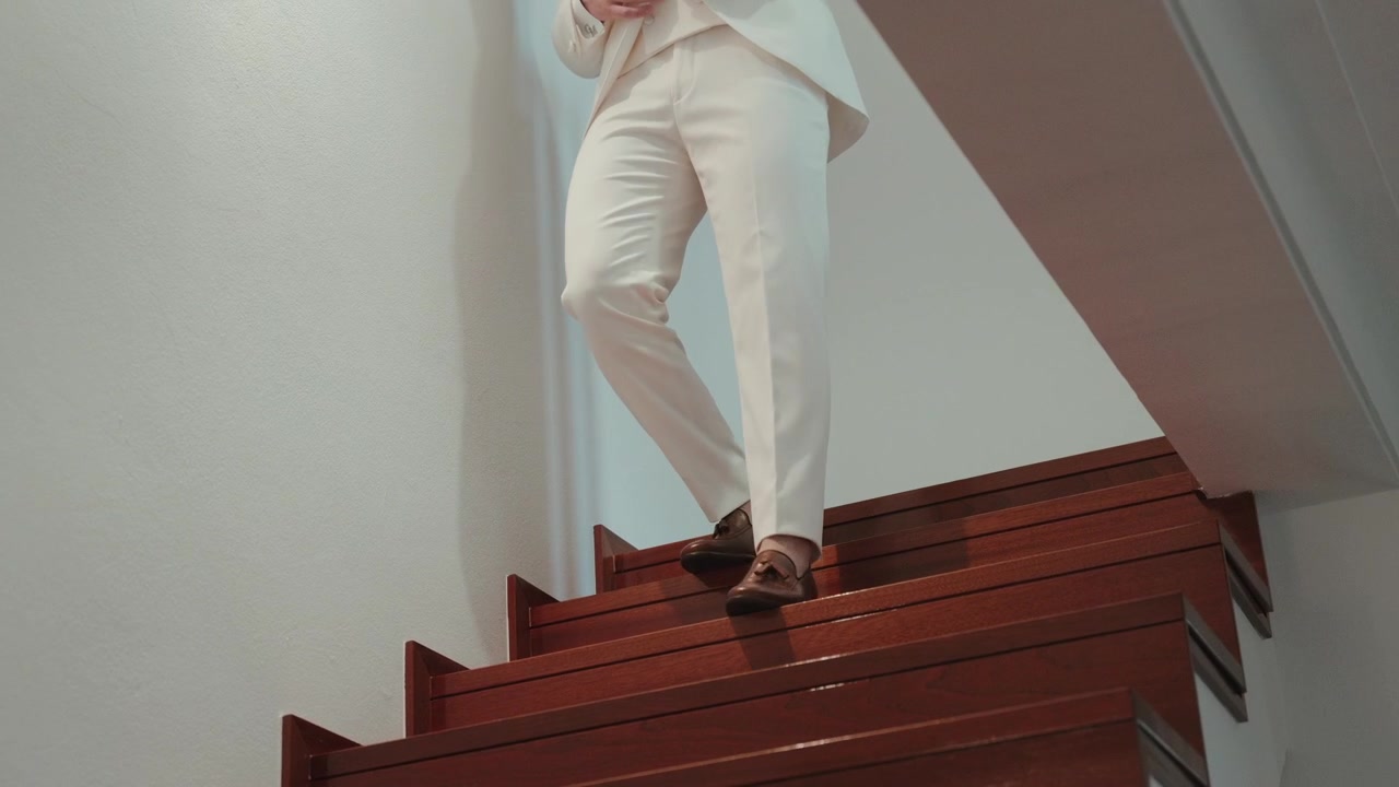 Groom in cream trousers and loafers descending wooden stairs indoors