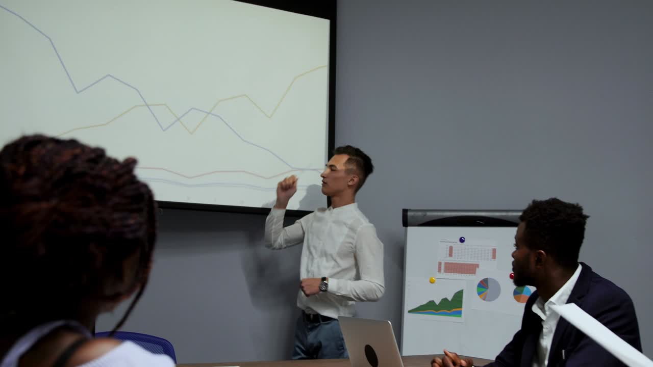 A young employee of the company in a white shirt reports on the progress of the work done over the past year and the development plans for the busy quarter on the diagram of the projector in the office. Report to the Board of Directors of a multi-ethnic