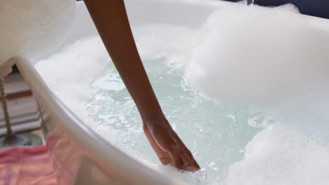 african american woman with towel preparing bath and touching water in bathroom