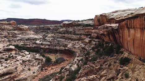 Drone-fly-along-rock-face-with-winding-riverbed-in-Utah-canyon