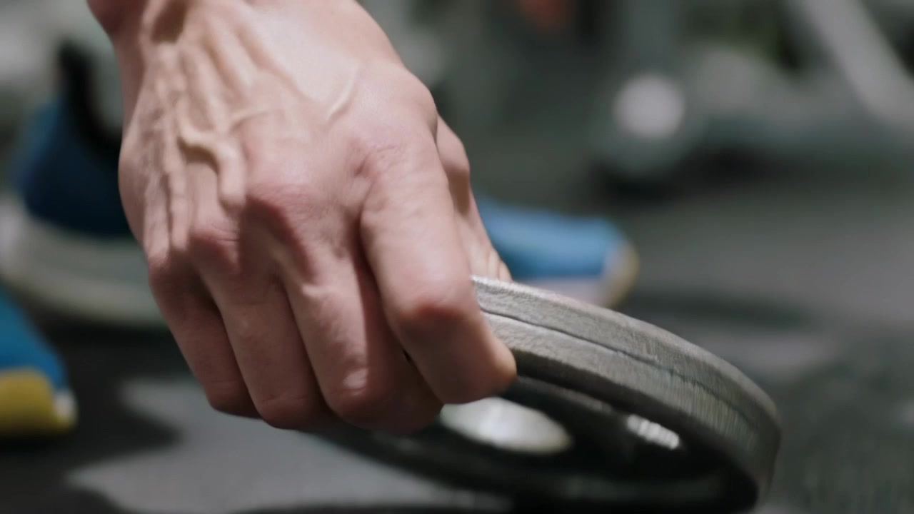 ジムで、タンクトップを着て青い靴を履いた男性が、カール運動の準備として、手を伸ばしてウェイトプレートをバーに装着している。