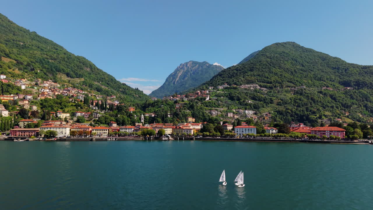 Drone flies backward over Lake Como, revealing two small sailboats, the coastal town, and the surrounding green mountains