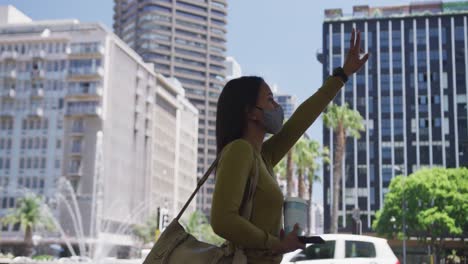 african american woman wearing face mask holding coffee raising hand for a taxi