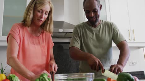 Happy-diverse-senior-couple-chopping-vegetables-in-kitchen-and-talking
