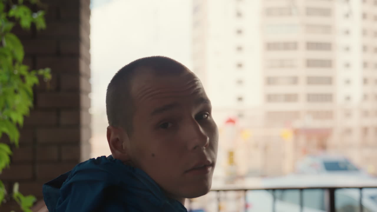 Cappuccio rimosso che rivela un ritratto contemplativo su un balcone, un giovane uomo si gira verso la camera con sguardo misurato, testa rasata ed espressione discreta, luce naturale soffusa e skyline della città sullo sfondo, pianta in vaso