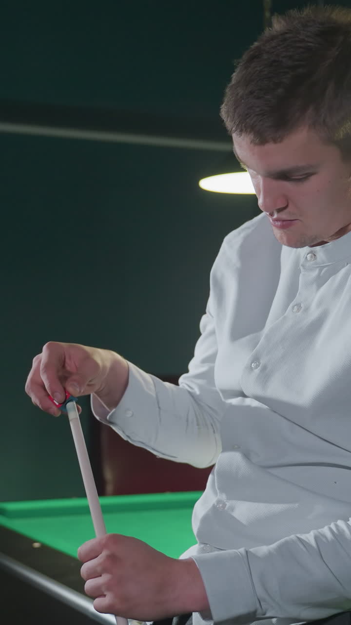 primer plano de un joven con camisa blanca, sentado, sosteniendo un palo de taco y aplicando cuidadosamente tiza roja con expresión enfocada. configuración de sala de billar débilmente iluminada con mesa de billar verde y bolas en el fondo