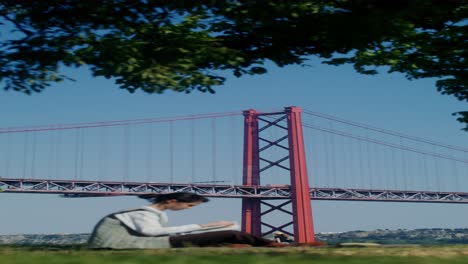 woman reading in a park with the 25 de abril bridge