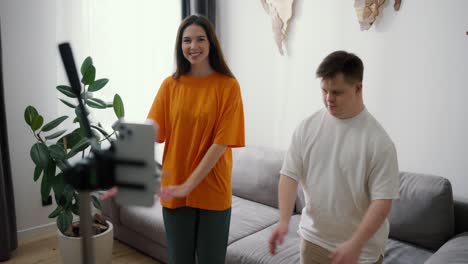 Un-Joven-Con-Síndrome-De-Down-Bailando-Frente-A-La-Cámara-Móvil-En-Casa-Con-Una-Chica-Sonriente-En-Pareja