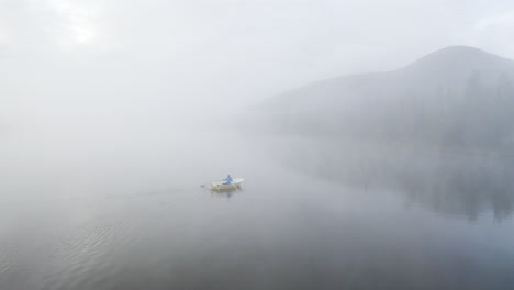 flying above a lake next to a small boat with a person rowing on a misty morning with mountain in the background