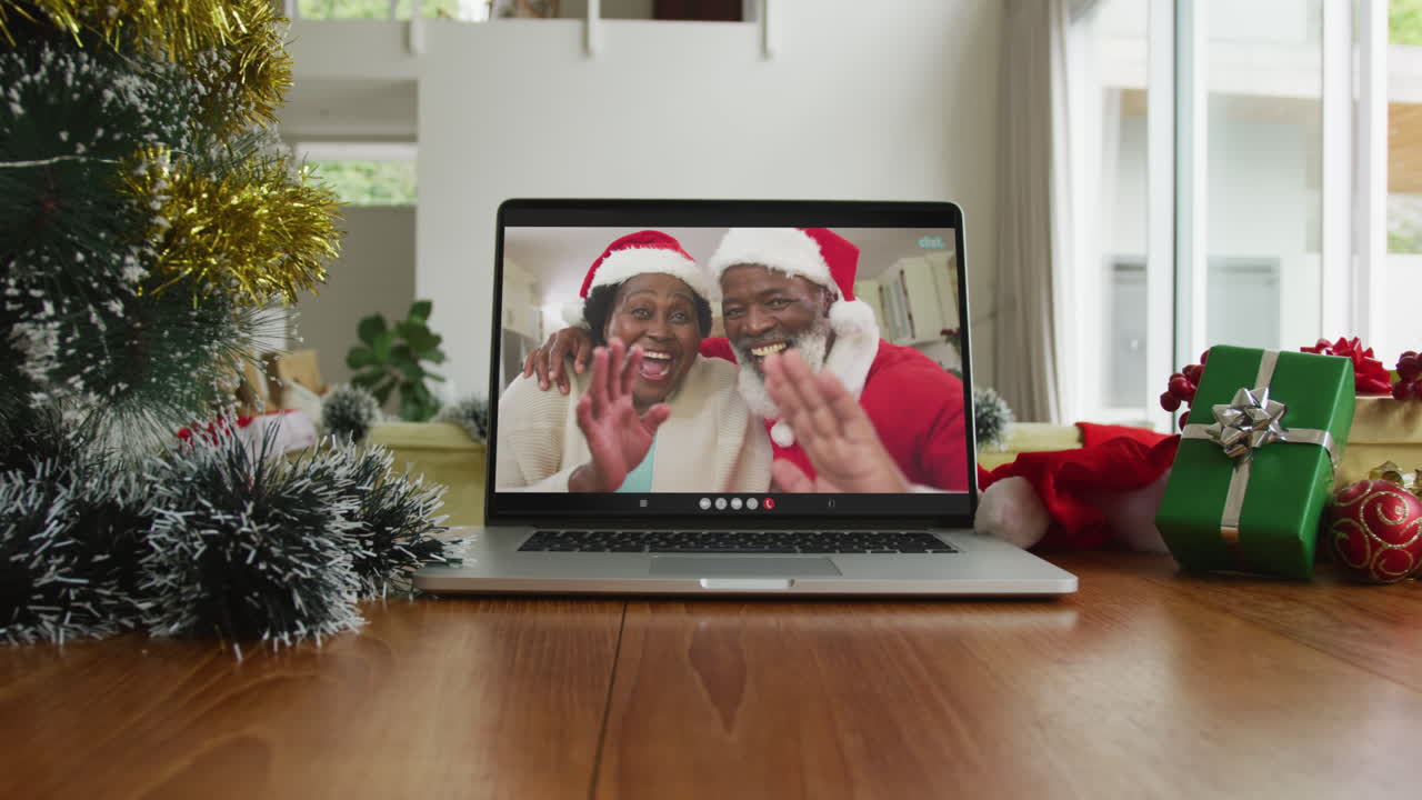 una pareja afroamericana sonriente agitando y usando sombreros de santa en la videollamada de navidad en una computadora portátil