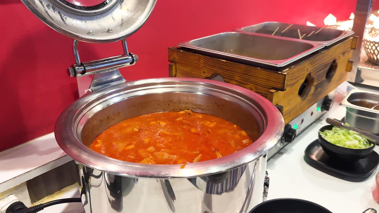 A person serves kimchi soup from a pot in a vibrant buffet setting with warm lighting