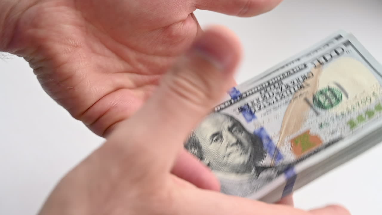 A man's hands quickly count a stack of one hundred dollar bills against a light background