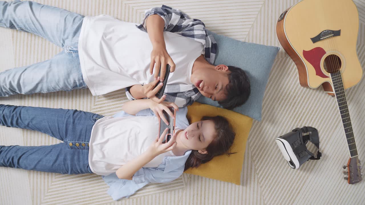 아시안 십대 커플이 집 바닥에 카에 누워있는 동안 스마트폰에서 함께 게임을 즐기고 있습니다.