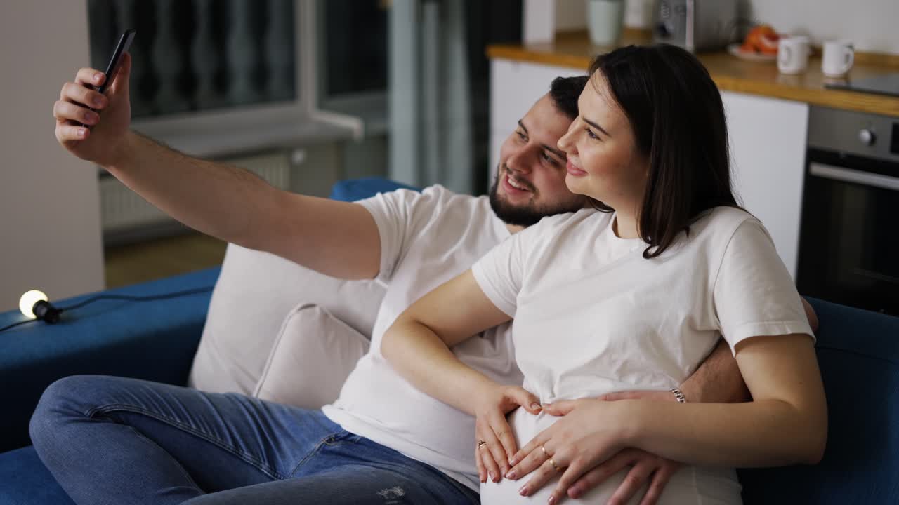 hombre feliz y su esposa embarazada tomando una selfie con un teléfono inteligente en casa