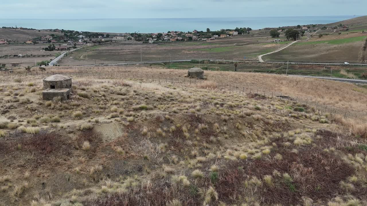 Roterende luftoptagelser viser flere bunkere fra Anden Verdenskrig spredt ud over en tør siciliansk mark, med Middelhavet synligt bag det bølgende terræn