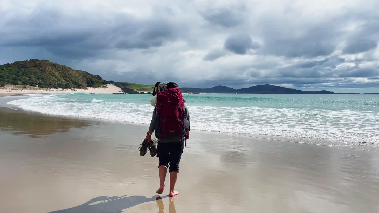 mujer mochilera camina sobre la arena y las olas para mirar las nubes oscuras y la vista del océano