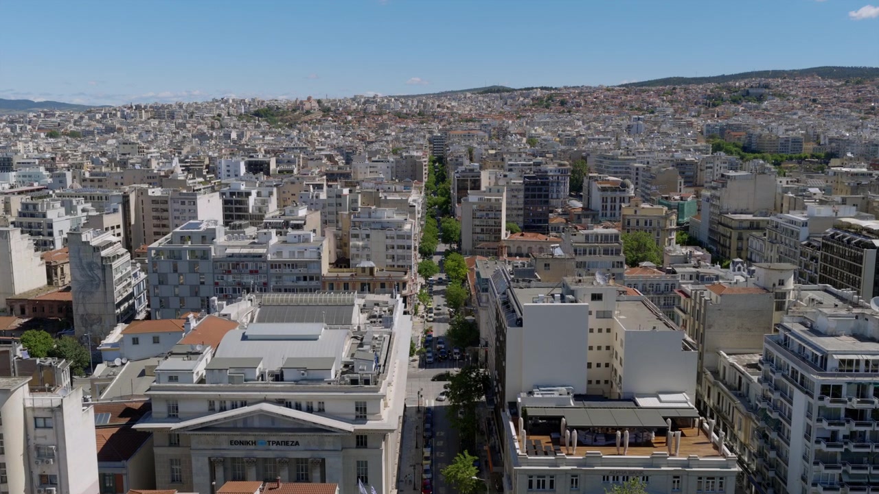 Aerial view down a tree-lined city avenue in Thessaloniki, flanked by historic buildings and dense hillside neighborhoods beyond.