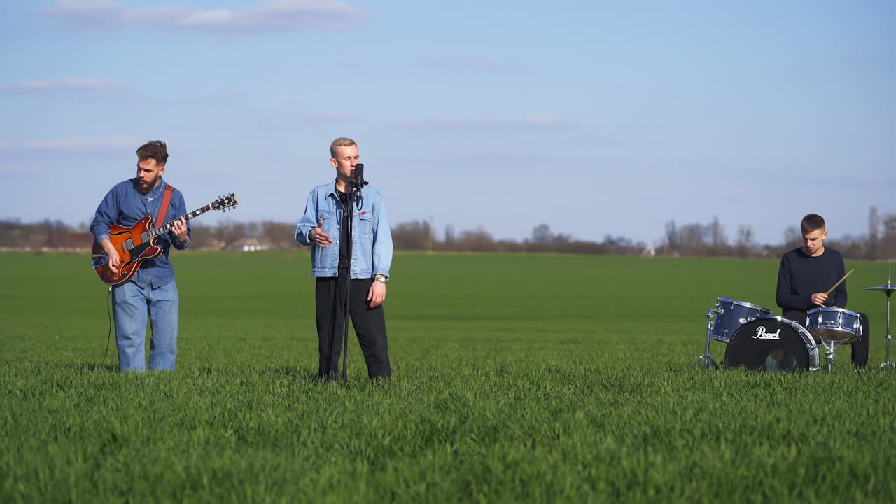 Guitarist, drummer and singer make performance among the fields. Young men musicians play music in the nature backdrop.