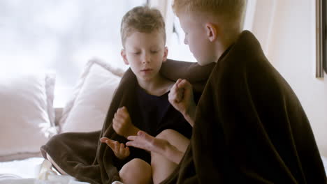 two little brothers with blanket on their shoulders sitting on bed in a campervan and playing rock scissors paper