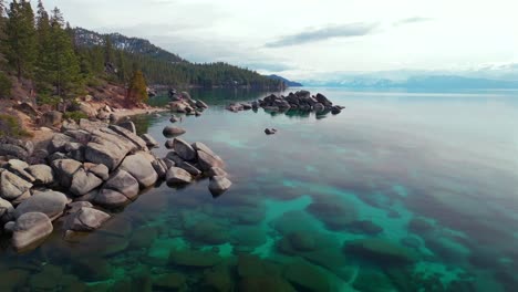 lake tahoe rocky shoreline large boulders crystal clear glassy turquoise fresh water aerial drone sand harbor california sunny daytime winter clouds secret chimney beach incline village forward pan up