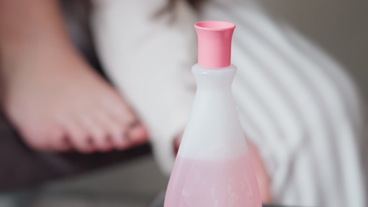 Female hand opens pink cap of nail remover bottle, sets cover aside while toes rest nearby, close view of careful grip and twist, soft indoor light, beauty care moment before pedicure cleanup