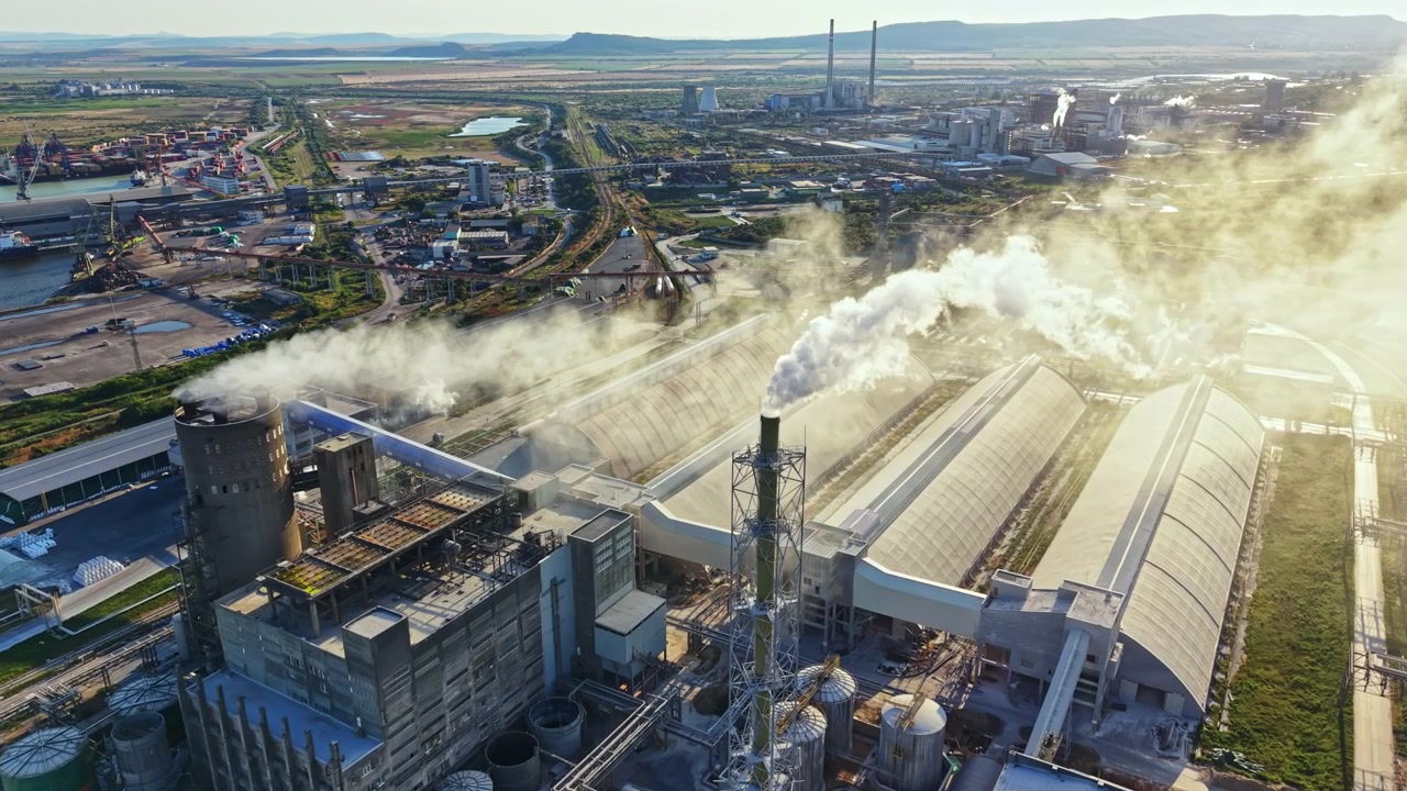 Il fumo si alza da edifici industriali in un'area di fabbriche durante il giorno, mentre veicoli si muovono su strade vicine e macchinari operano sullo sfondo