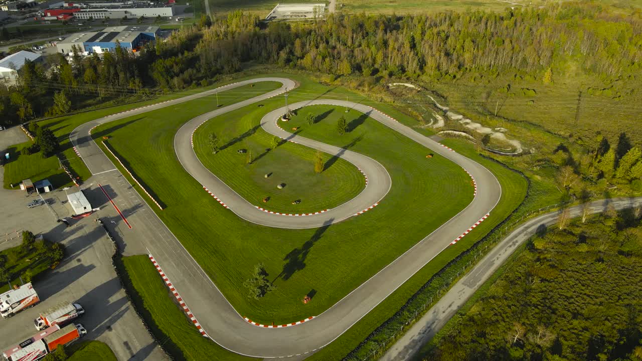 Aerial drone footage orbiting and flying around a go kart or karting racing speedway track in a sunny summer or autumn countryside. The track is in the middle of a forest and besides a highway road