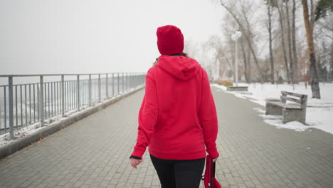 back view of woman wearing red jacket walking along interlocked pathway holding bag in left hand near iron railing in serene park with snowy ground, benches, and trees in foggy winter atmosphere