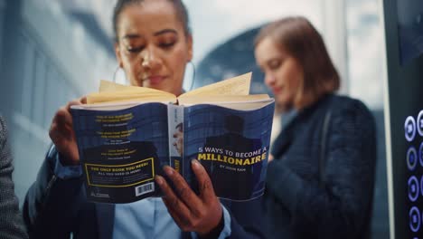 confident black adult female riding glass elevator to office in modern business center. african american manager reading a finance book called "5 ways to become millionaire" in a lift.