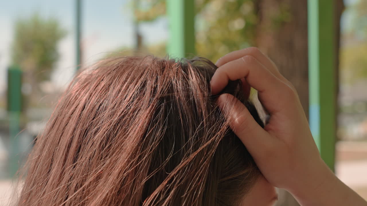 정류장에서 머리를 매만지는 손의 클로즈업, 햇빛에 비친 갈색 머리카락이 산들바람에 흩날리고, 부드럽게 두피를 만지며 스타일링하는 모습, 교외 배경과 편안한 정오의 햇살 아래 자연스러운 통근자의 순간