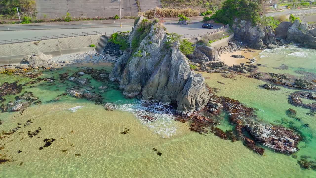 An aerial shot circles a large, tree-topped rock formation in the clear turquoise sea, revealing Japan's beautiful coastline and a nearby coastal road