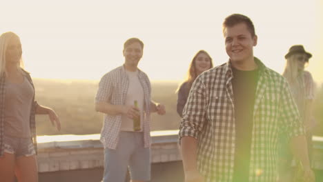 a young man in a gray plaid shirt and black t-shirt moves in dance with his friends. he enjoys and has fun with beer and music on the roof.