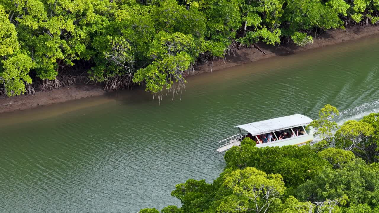 Aerial footage of a tour boat carrying passengers along a winding river bordered by dense rainforest, under bright daylight with steady drone movement