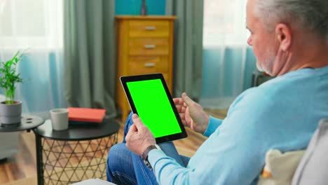 el hombre mayor en casa usa una tableta de pantalla de maqueta verde. está sentado en un sofá en su acogedora sala de estar. el hombre hace clic una vez en la pantalla y navega por el contenido. por encima del hombro de la cámara disparada