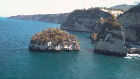 Aerial-view-of-the-crystal-blue-water-of-diamond-beach-in-Nusa-Penida,-Indonesia-with-a-prominent-large-cliff-in-the-foreground