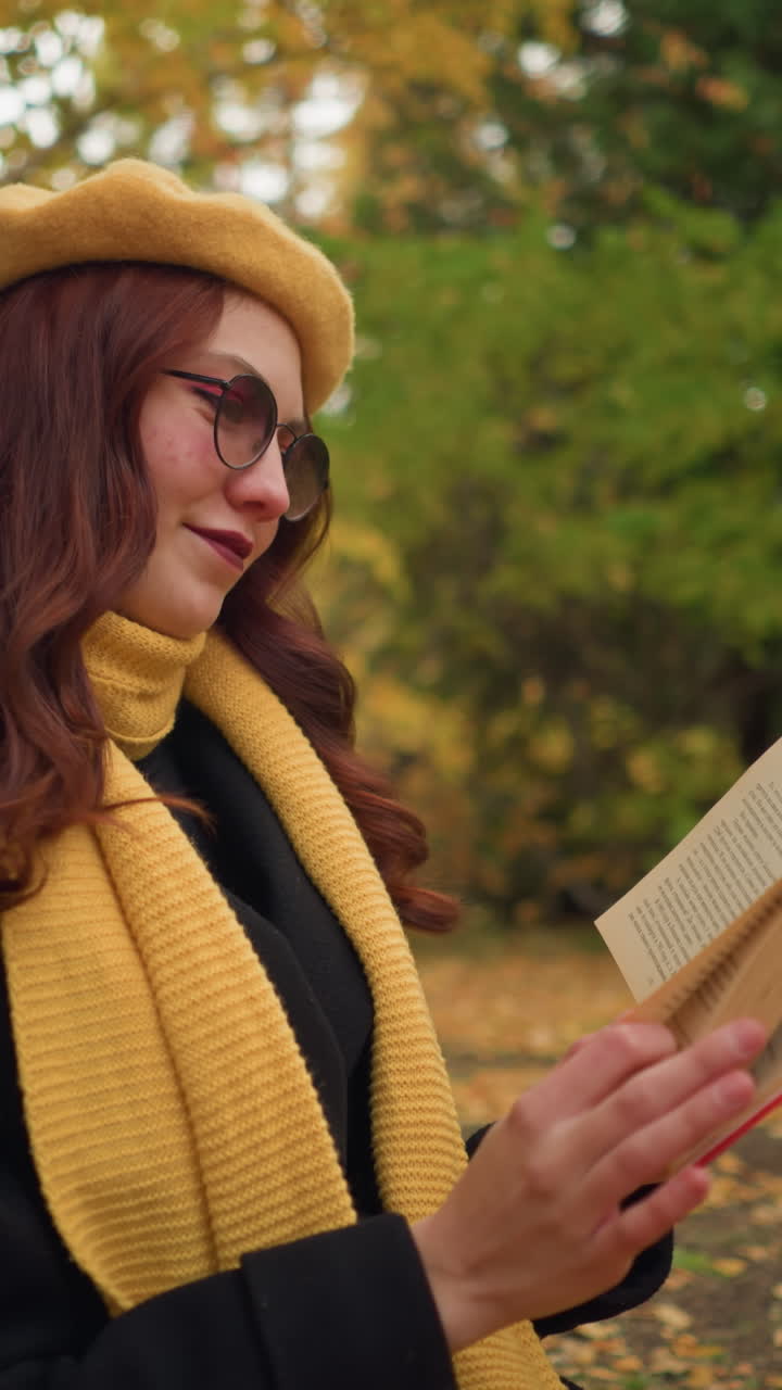 estudiante en la soledad lee a través de libro cubierto amarillo con sonrisa cálida, sentado solo en la naturaleza, con boina amarilla y silenciador amarillo, apoyado en el abedul rodeado de hojas de otoño